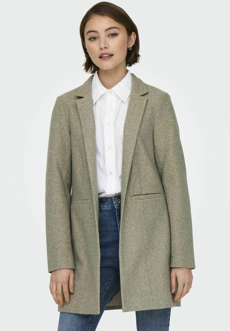 Cappotto in misto lana verde chiaro con design su misura, maniche lunghe, due tasche laterali e chiusura frontale con un bottone sopra una camicia bianca.