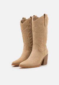 Botas de vaquero de ante beige con punteras puntiagudas, patrones intrincados de costura y un tacón apilado. Cuentan con tiradores en la caña para facilitarel uso.