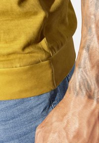 Tattooed forearm next to torso wearing a yellow top and blue denim jeans, shown in close-up with fabric texture visible.