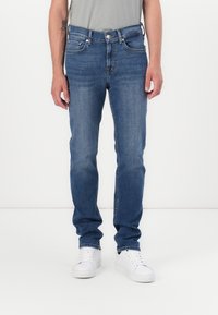 Jean en denim bleu à coupe droite, avec cinq poches et des accessoires métalliques. Porté avec des baskets blanches et un T-shirt gris.