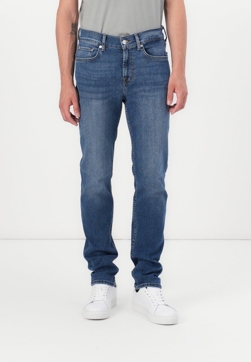 Jean en denim bleu à coupe droite, avec cinq poches et des accessoires métalliques. Porté avec des baskets blanches et un T-shirt gris.