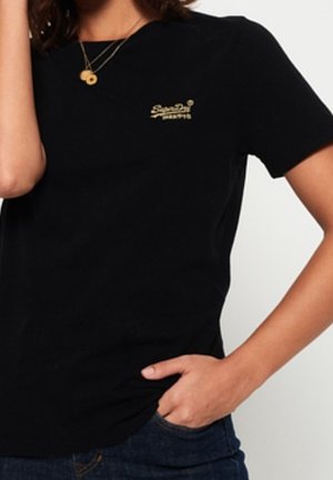 Person wearing a black Superdry t-shirt with small gold logo on chest, hand in blue jeans pocket, and layered gold necklaces.