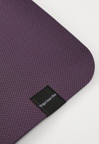 Fialová joga podložka s textúrovaným povrchom, zaoblenými rohmi a čiernou nálepkou s nápisom "Yoga Searcher". Odolný, protišmykový materiál.