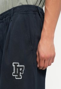 Námornícke jogger nohavice vyrobené z ľahkého materiálu, s elastickým pásom a bočnými vreckami. Vyšitý logo v sivej farbe na stehne.