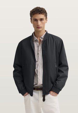 Giovane uomo con giacca nera con zip sopra una camicia a righe marroni e pantaloni bianchi, in piedi con le mani in tasca davanti a uno sfondo semplice.