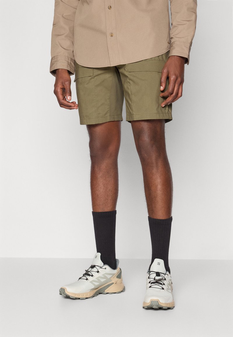Pantaloni cargo color khaki con tasche frontali piatte, abbinati a sneakers bianche e beige e calzini neri a costine, indossati con una camicia beige con bottoni.