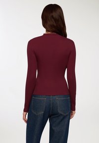 Top lungo bordeaux a maniche lunghe con design aderente, texture a coste e scollatura rotonda, abbinato a jeans in denim blu a vita alta.