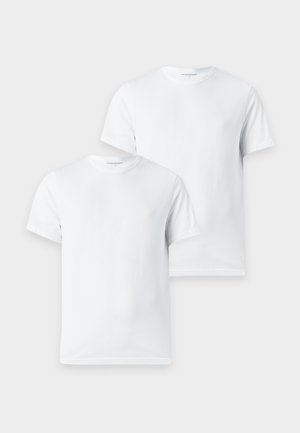 Mann steht barfuß, trägt ein schlichtes weißes Kurzarmshirt und schwarze enganliegende Shorts vor einem schlichten hellen Hintergrund.