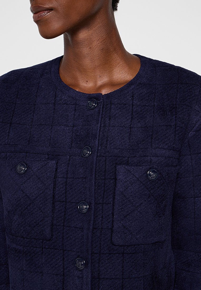 Granatowa kurtka z fakturą, w kratę, okrągłym dekoltem i sześcioma czarnymi guzikami. Zawiera dwie przednie kieszenie naszywane.