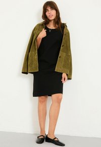 Olivengrüne Wildlederjacke über einem schwarzen Kleid mit kurzen Ärmeln und geradem Schnitt. Schwarze flache Schuhe runden das Outfit ab. Schlichte Gestaltung.