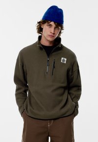 Olívovo zelený pulóver s textúrovan�ým vzorom, polovičným zipsovým golierom a čiernymi zipsami. Na hrudi má nášivku s logom a je nosený s hnedými nohavicami.