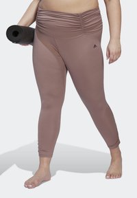 Bruine rekbare leggings met een verzamelde tailleband, gemaakt van soepel materiaal, gedragen met een zwarte yogamat in de hand, op een lichte achtergrond.