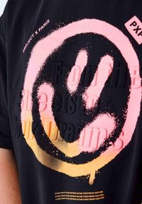 Camiseta negra con un gran gráfico de cara sonriente rosa con efecto de degradado. El texto dice "De las calles a tus sueños" en colores superpuestos.