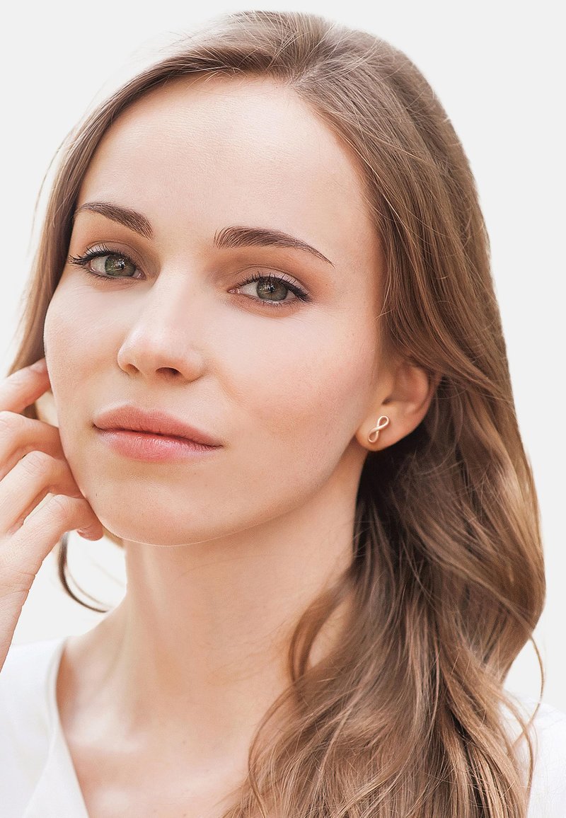 Goldfarbene Ohrringe in Form eines stilisierten Unendlichkeitssymbols, getragen von einer Person mit langen, welligen braunen Haaren und natürlichem Makeup.