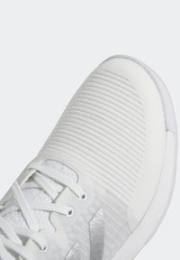 Vit sportskor med en texturerad ovandel med horisontella ribbor, snörning och lätt dämpning. Subtila reflekterande detaljer finns.