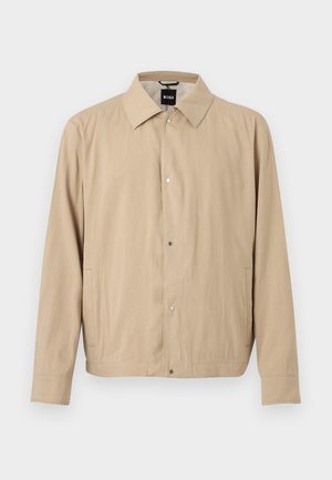 Μπεζ ελαφρύ μπουφάν με μακριά μανίκια, κουμπιά με πάτημα μπροστά, πλαϊνές τσέπες και κλασικό γιακά, κρεμασμένο σε λευκό φόντο.