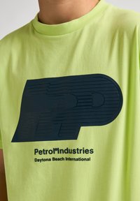 Camiseta de algodón verde neón con un gran gráfico negro con líneas y el texto "Petrol Industries Daytona Beach International".