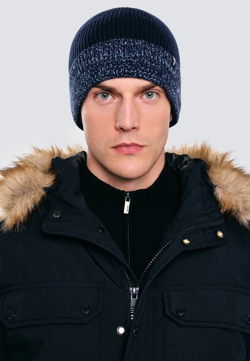 Granatowa dzianinowa czapka z teksturowanym wzorem na szczycie. W połączeniu z czarną kurtką z kapturem obszytym futerkiem i zamkiem błyskawicznym z przodu.
