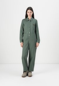 Combinaison verte à manches longues, fermeture à boutons, poches poitrine et coupe décontractée. Fabriquée en tissu texturé, portée avec des chaussures beiges.