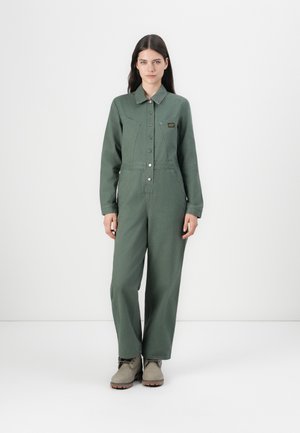 Green jumpsuit with long sleeves, button closure, chest pockets, and a relaxed fit. Made of textured fabric, worn with beige shoes.