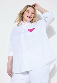 Hvid oversized skjorte med knaplukning og et pink hjertepatch på venstre lomme. Blødt stof, opsmøjede ærmer og en buet nederkant.