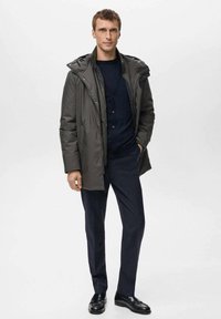 Cappotto imbottito grigio scuro con cappuccio, indossato sopra un maglione blu navy, abbinato a un paio di pantaloni scuri e scarpe nere lucide. Design semplice e funzionale.