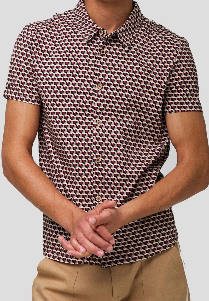 Chemise à manches courtes boutonnée avec un motif géométrique de cubes en rouge, noir et beige, portée avec un pantalon beige, mains jointes devant.