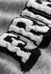 Sweter z dzianiny w szarym kolorze, z wyrazistymi czarnymi i białymi wzorowanymi literami. Tekstura wydaje się być miękka z nieco wypukłym wzorem.