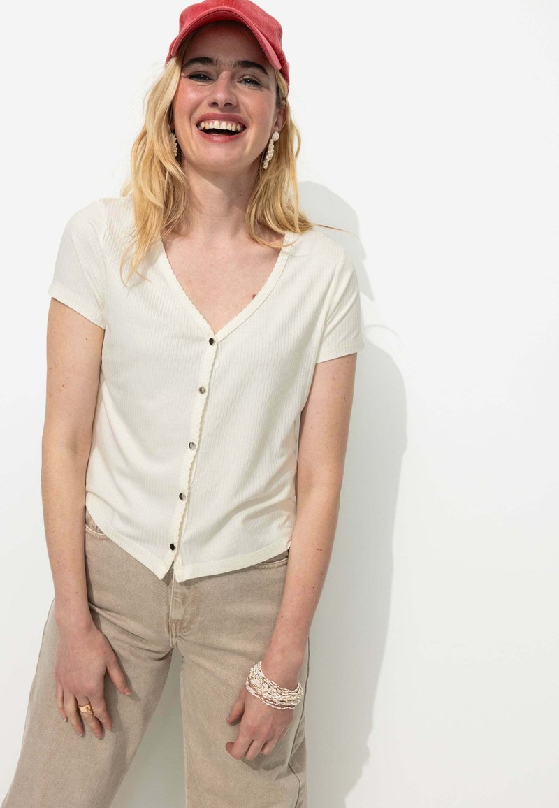 Top crème à manches courtes côtelé avec un décolleté en V et une fermeture à boutons. Pantalons larges couleur marron clair. Les accessoires incluent un bracelet superposé.