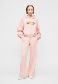 Ροζ βαμβακερό φούτερ με κουκούλα και μπροστινό γραφικό κείμενο σε διάφορα χρώματα, σε συνδυασμό με ροζ παντελόνια άνετης εφαρμογής και λευκά παπούτσια.