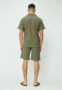 Chemise côtelée à manches courtes vert olive et shorts assortis, offrant une coupe décontractée et une texture lisse. Des tongs noires complètent le look.