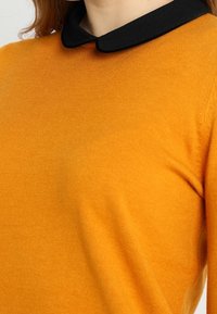 Pull jaune moutarde avec un col noir, fabriqué en tissu doux et texturé. Présente une coupe ajustée, montrant un gros plan sur l'encolure.