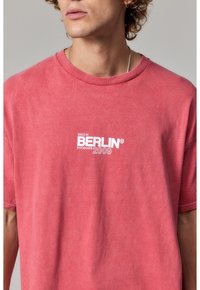 Czerwona, krótkosleeve koszulka wykonana z bawełny o luźnym kroju. Posiada białe nadrukowane napisy: „ISSUE 01 BERLIN POTSDAMER 2009.” Tekst jest wyśrodkowany.