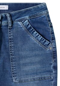 Poche de jean en denim bleu avec coutures visibles, passant de ceinture et détail de rivet métallique sur la partie avant supérieure.