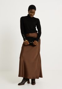 Jersey negro de cuello alto con acentos de perlas en los puños, combinado con una larga falda de satén marrón y un bolso negro de mano con un asa circular.