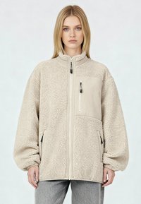 Giacca in pile beige con zip frontale, collo alto e grandi tasche. Caratterizzata da una texture morbida e da una tasca sul petto a contrasto con cerniera.