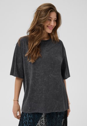 Kvinde med langt brunt hår smiler, iført en løs sort vasket T-shirt og en mørk blondeskørt, står mod en ensfarvet lys baggrund.
