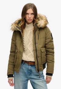 Oliwkowa kurtka puchowa z kapturem z sztucznego futra, z zamkiem błyskawicznym, bocznymi kieszeniami i ściągaczami przy rękawach. Noszona na kremowym swetrze w warkocze i niebieskich dżinsach.