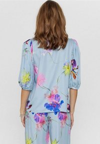 Light blue blouse with puff sleeves, featuring a floral pattern in pink, purple, and yellow hues. Smooth fabric with a relaxed fit.