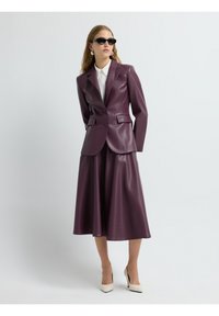 Blazer in ecopelle viola e gonna midi coordinata, con un design sartoriale e una texture liscia, abbinati a una camicia bianca e tacchi.