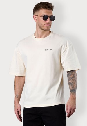Cremfarbener Baumwoll-T-Shirt mit kurzen Ärmeln, seitlichen Schlitzen und einem kleinen schwarzen Logo auf der Brust. Model trägt schwarze Sonnenbrillen.