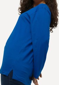 Haut à manches longues bleu en tissu doux, doté d'un décolleté en V et de fentes sur les côtés pour faciliter les mouvements. Texture lisse et coupe décontractée.