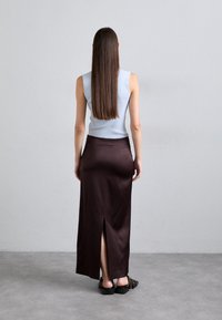 Fustă din satin maro cu slit la spate, asortată cu un top fără mâneci de culoare albastru deschis. Păr lung și drept vizibil, stând pe un podea gri.