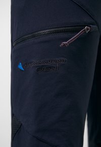 Gros plan d'un pantalon en tissu noir avec poche zippée, logo triangle bleu brodé et texte noir indiquant "Blåhornmusen Hærmod."