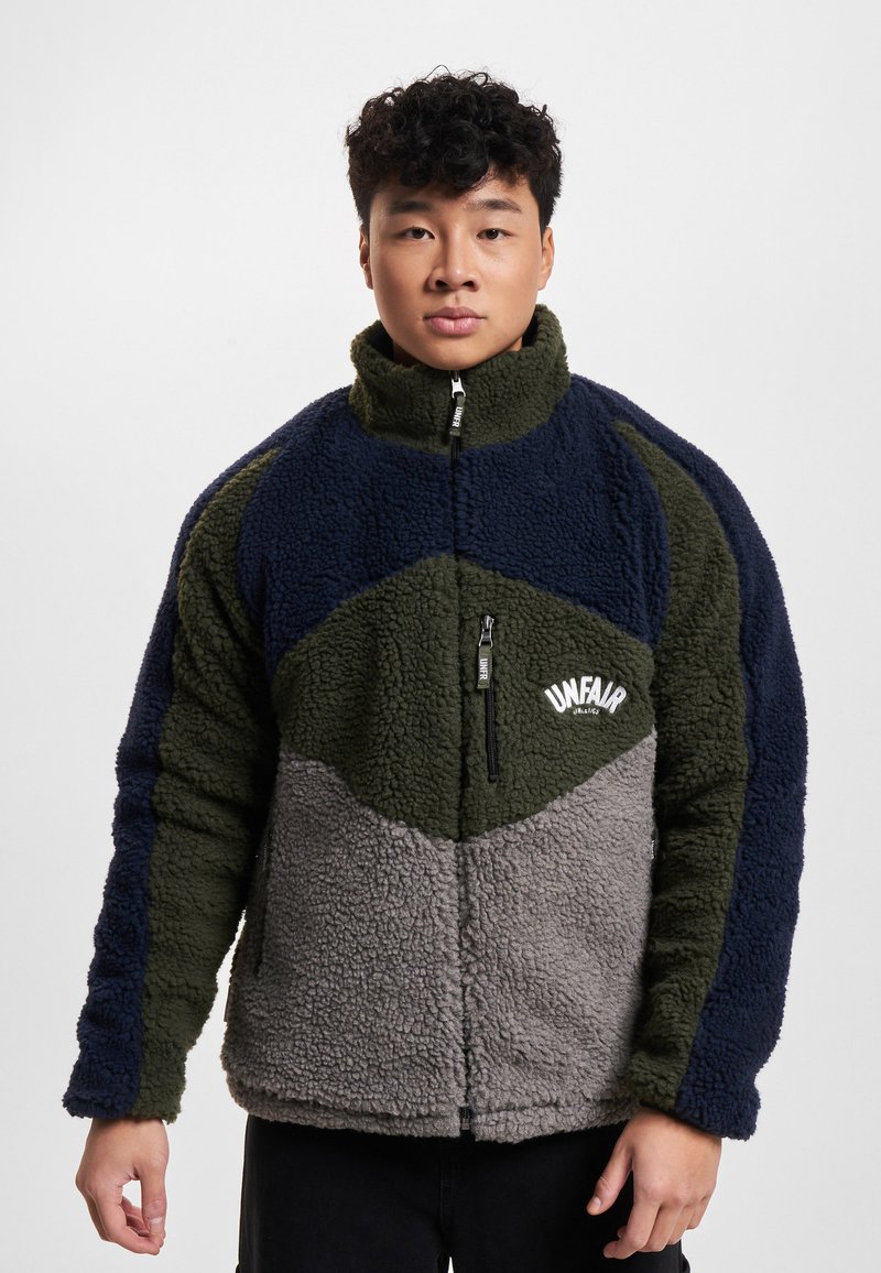 Jeune homme portant une veste polaire bicolore bleu marine, vert olive et gris avec une fermeture éclair à l'avant et le logo "UNFAIR" sur la poitrine.