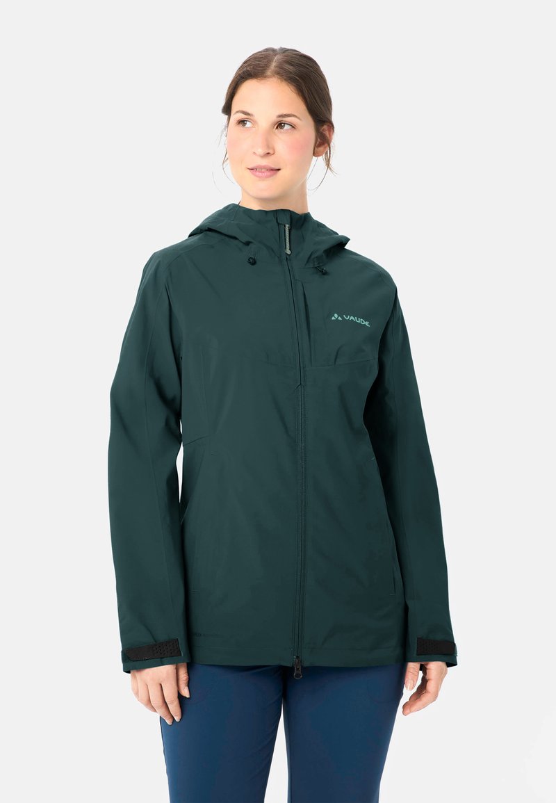 Frau trägt eine dunkelgrüne Kapuzenjacke mit Reißverschluss und Logo, steht mit entspannten Händen und schaut leicht nach links.