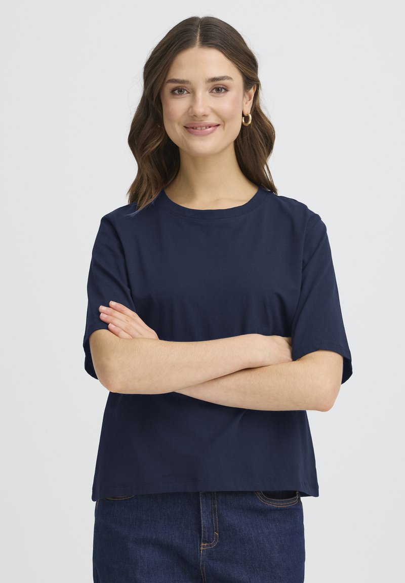 Temno modra bombažna majica z okroglim izrezom in kratkimi rokavi, z rahlo ohlapnim krojem in gladkim teksturo, kombinirana s temnimi jeans hlačami.