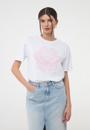 T-shirt bianco in cotone con una grande grafica di una farfalla rosa e elementi floreali. Abbinato a una gonna di jeans blu chiaro.