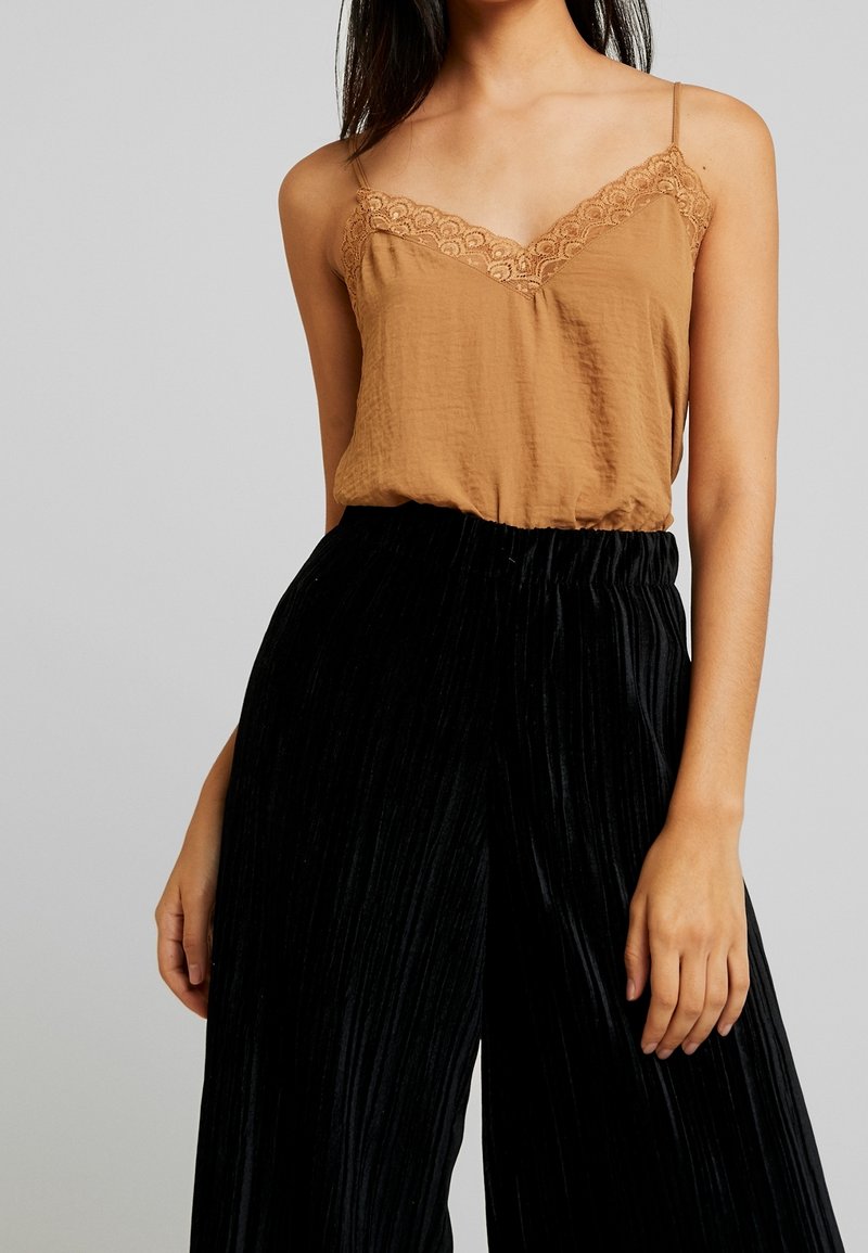 Femme portant un haut marron sans manches avec bordure en dentelle et un pantalon noir plissé taille haute, visible du cou à la mi-cuisse.