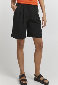 Schwarze, maßgeschneiderte Shorts mit Falten, Seitentaschen und glatter Textur, kombiniert mit schwarzen Sandalen. Das Hemd ist orange und sorgt für Kontrast.